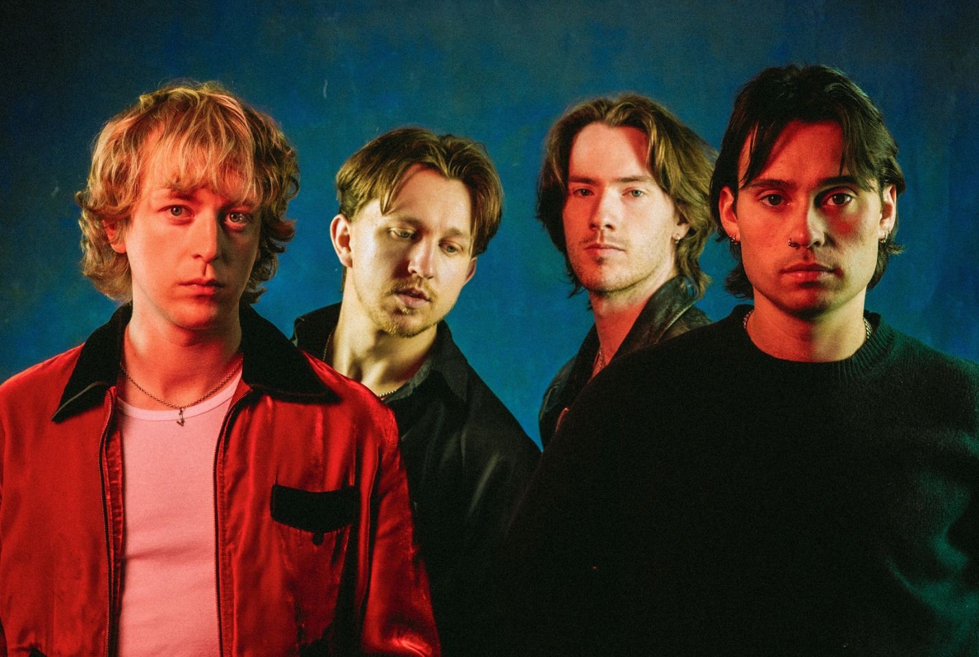 The four band members of Balancing Act stood in front of a blue background, with a red light shining towards them. Three are wearing black jackets, one is wearing a red one.